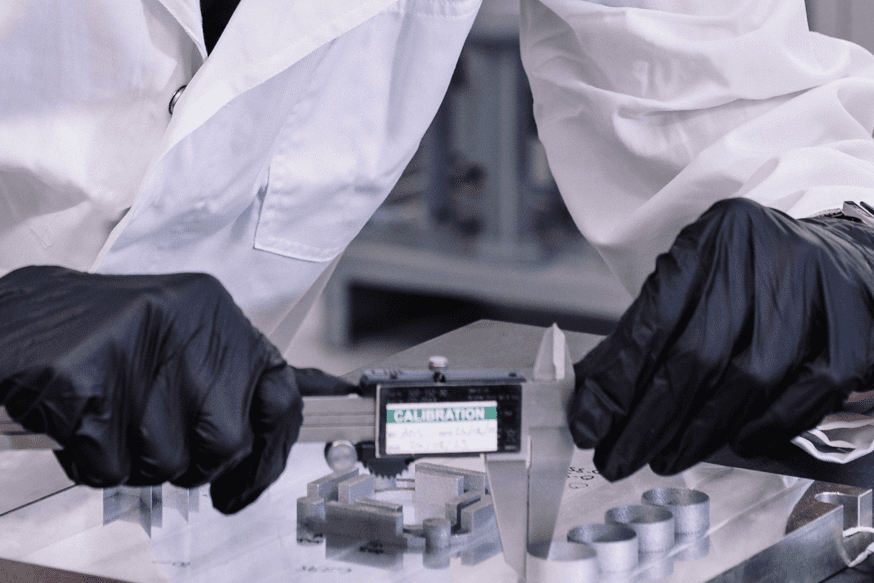 close up of hands in black gloves measuring a metal AM part with a pair of vernier calipers